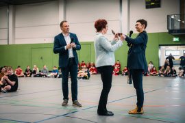 Drei Personen stehen in der Mitte der neuen Drei-Feld-Sporthalle. Frau Hofmann-Dubrau von der Stadt Hoyerswerda übergibt der Schulleiterin Romy der Oberschule Hoyerswerda, Romy Stötzner, einen großen symbolischen Schlüssel für die neue Sporthalle. Links neben ihnen steht Bürgermeister Mirko Pink und applaudiert. Ringsherum sitzen Schüler.