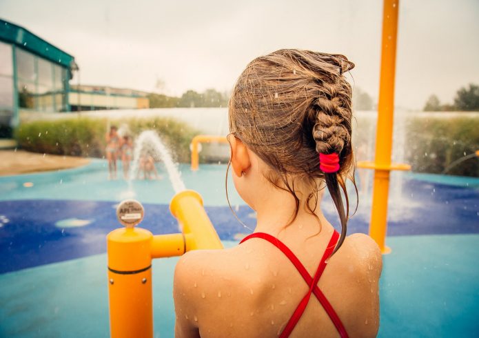 Ein Mädchen mit geflochtenem Zopf und rotem Badeanzug steht im Außenbereich des Lausitzbads Hoyerswerda an einem gelben Wasserspielgerät und steuert einen Wasserstrahl. Im Hintergrund sind weitere Kinder in Badebekleidung zu sehen, die von anderen Wasserdüsen bespritzt werden. Der Boden ist mit rutschfestem, blauem Belag versehen, rundherum sind Büsche und Gebäude zu erkennen. Die Szene wirkt lebendig und sommerlich.