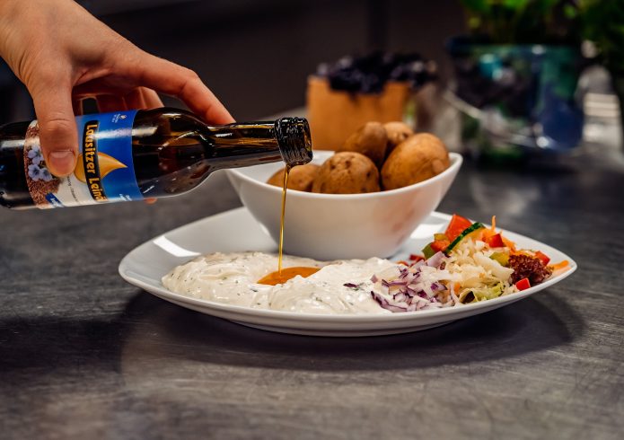 Ein Teller mit regionaler Speise im Gastrobereich des Lausitzbades Hoyerswerda. Im Vordergrund wird aus einer Flasche „Lausitzer Leinöl“ ein goldgelber Strahl Öl über einen Dip aus Quark mit Kräutern gegossen. Auf dem Teller liegen zudem ein bunter Salat mit roten Zwiebeln und eine Schale mit gekochten Pellkartoffeln. Im Hintergrund sind Küchenutensilien und frische Kräuter zu sehen.