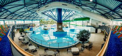Innenaufnahme des Lausitzbades Hoyerswerda: In einem großen Erlebnisbecken mit blauen Fliesen schwimmen bunte Reifen – rot, gelb und blau – in einem separaten Rundbecken. Rechts im Hintergrund mündet eine grüne Wasserrutsche ins Becken. Über dem Wasserbecken befindet sich eine Plattform mit einer Deko-Figurengruppe: drei Affen, die nach dem Motto „nichts sehen, nichts hören, nichts sagen“ posieren. Palmen und tropische Pflanzen schaffen eine spielerische Atmosphäre in der hellen, lichtdurchfluteten Badehalle.