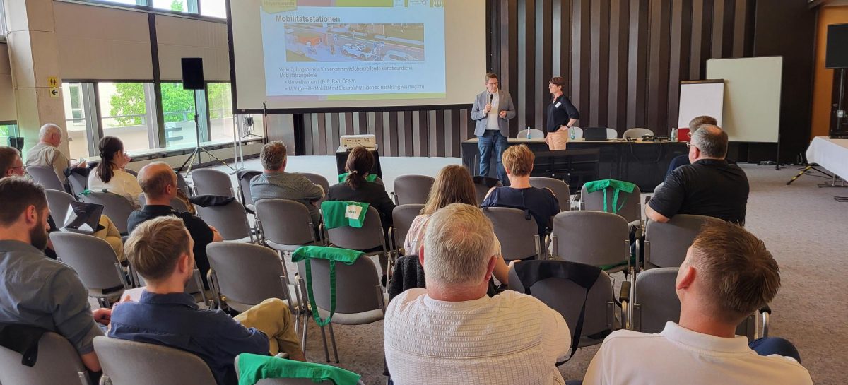 In einem Konferenzraum sitzen mehrere Personen auf Stühlen und verfolgen eine Präsentation. Auf einer großen Leinwand vorn ist eine Folie mit dem Titel „Mobilitätsstationen“ zu sehen, die sich mit multimodaler Mobilität beschäftigt. Zwei Vortragende – ein Mann mit Mikrofon und eine Frau – stehen vorn und sprechen. Die Atmosphäre ist konzentriert, das Licht fällt durch große Fenster in den Raum.