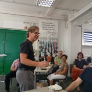 Ein Mann mittleren Alters mit Brille und Headset-Mikrofon steht lächelnd vor einer Gruppe sitzender Personen in einem Seminarraum. Er hält eine Kaffeetasse in der Hand. Im Hintergrund ist ein Plakat mit Binärcode und der Aufschrift „Wenn Computer miteinander reden, welche Sprache sprechen sie dann?“ zu sehen. Einige Zuhörerinnen und Zuhörer blicken interessiert zum Redner.