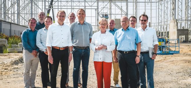 Eine Gruppe von elf Personen steht vor einer großen Stahlkonstruktion auf einer Baustelle. In der Mitte Staatsministerin Regina Kraushaar und Oberbürgermeister Torsten Ruban-Zeh. Alle tragen sommerliche Kleidung, keine Schutzhelme.