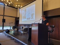 Eine Frau im schwarzen Oberteil steht am Rednerpult im Forumsaal in der Lausitzhalle Hoyerswerda und spricht in ein Mikrofon. Auf der Leinwand hinter ihr ist eine Präsentationsfolie mit historischen Abbildungen und dem Titel „Das 1000-Mann-Lager“ zu sehen.