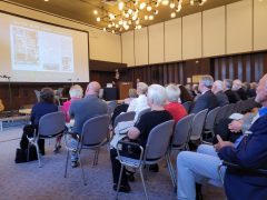 Ein Saal voller Menschen sitzt in Reihen und verfolgt einen Vortrag. Vorn am Rednerpult steht eine Frau und spricht in ein Mikrofon. Auf der Leinwand hinter ihr ist eine Präsentationsfolie mit dem Titel „Das 1000-Mann-Lager“ und historischen Abbildungen zu sehen.