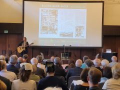 Eine Frau steht auf einer Bühne in der Lausitzhalle Hoyerswerda und spielt Gitarre. Sie singt in ein Mikrofon, während hinter ihr eine große Leinwand eine Präsentationsfolie mit der Überschrift „Das 1000-Mann-Lager, Hoyerswerda 1955–1970“ zeigt. Im Vordergrund sitzt das Publikum und hört zu.