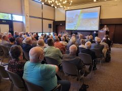 Ein Mann im grauen Anzug spricht am Rednerpult in der Lausitzhalle Hoyerswerda. Auf der Leinwand hinter ihm ist eine Präsentation mit historischen und aktuellen Bildern der Stadt zu sehen, überschrieben mit „70 Jahre Hoyerswerda-Neustadt – Herzlich Willkommen!“. Vor ihm sitzt ein vollbesetzter Saal mit Gästen, die seiner Abschlussrede zuhören.