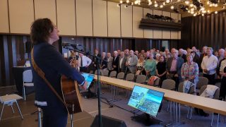 Eine Frau mit Gitarre steht auf der Bühne und singt in ein Mikrofon. Vor ihr stehen zahlreiche Menschen im Saal, die gemeinsam mitsingen. Es handelt sich um den Moment, in dem das traditionelle Steigerlied bei der Festveranstaltung zum 70-jährigen Jubiläum der Hoyerswerdaer Neustadt angestimmt wird.