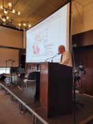 Ein älterer Mann im weißen Hemd spricht am Rednerpult im Forumsaal der Lausitzhalle Hoyerswerda. Hinter ihm ist eine große Leinwand mit einer Karte zu sehen, die die Aufteilung Deutschlands nach dem Potsdamer Abkommen 1945 zeigt.