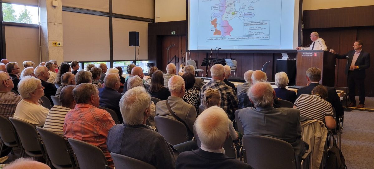 Ein älterer Mann im weißen Hemd spricht am Rednerpult in einem Saal voller Zuhörerinnen und Zuhörer. Auf der Leinwand ist eine Karte zu sehen mit dem Titel „Deutschland nach dem Potsdamer Abkommen 1945“. Das Publikum verfolgt aufmerksam den Vortrag.