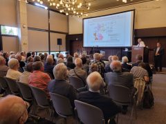 Ein älterer Mann im weißen Hemd spricht am Rednerpult in einem Saal voller Zuhörerinnen und Zuhörer. Auf der Leinwand ist eine Karte zu sehen mit dem Titel „Deutschland nach dem Potsdamer Abkommen 1945“. Das Publikum verfolgt aufmerksam den Vortrag.