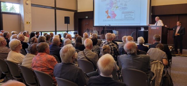 Ein älterer Mann im weißen Hemd spricht am Rednerpult in einem Saal voller Zuhörerinnen und Zuhörer. Auf der Leinwand ist eine Karte zu sehen mit dem Titel „Deutschland nach dem Potsdamer Abkommen 1945“. Das Publikum verfolgt aufmerksam den Vortrag.