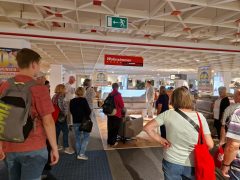 Eine Besuchergruppe steht in einem Möbelhaus im Bereich „Wohnzimmer“. Vor ihnen erklärt ein Mitarbeiter in hellem Anzug etwas zu den ausgestellten Sofas. Im Vordergrund und am Rand sind Teilnehmende mit Rucksäcken und roten Stoffbeuteln zu sehen.