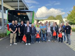 Eine Gruppe von rund 20 Teilnehmenden der „Hoyerswerdaer Spätschicht“ steht vor einem grünen Bus mit der Anzeige „VGH Spätschicht Tour 3“. Mehrere Personen halten rote Stoffbeutel in der Hand. Im Hintergrund sind Gebäude, Bäume und blauer Himmel mit Wolken zu sehen.
