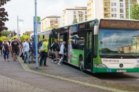 Teilnehmende steigen vor einem Wohnblock in Hoyerswerda in einen grünen Linienbus mit der Aufschrift „VGH Spätschicht Tour 2“ ein. Viele Menschen sind unterwegs, einige tragen Taschen.