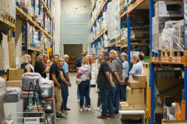 Eine Besuchergruppe läuft durch ein Hochregallager mit vollen Regalen. Ein Mitarbeiter führt sie und erklärt die Abläufe. Die Gruppe blickt interessiert nach oben.
