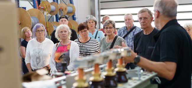 Eine Gruppe von Besucherinnen und Besuchern steht in einer Werkhalle vor Kabelrollen. Ein Mitarbeiter erklärt technische Geräte im Vordergrund. Alle hören aufmerksam zu.