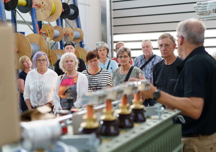 Eine Gruppe von Besucherinnen und Besuchern steht in einer Werkhalle vor Kabelrollen. Ein Mitarbeiter erklärt technische Geräte im Vordergrund. Alle hören aufmerksam zu.
