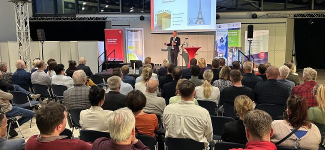 Heiko Lachmann steht auf einer Bühne vor einer Leinwand und spricht zu rund hundert Teilnehmerinnen und Teilnehmern des Unternehmerabends in Hoyerswerda. Auf der Leinwand ist eine Präsentationsfolie mit einer Grafik und dem Eiffelturm zu sehen.