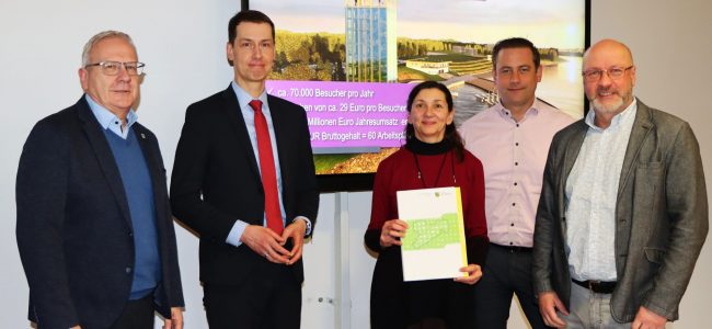 Fünf Personen stehen nebeneinander in einem Besprechungsraum. In der Mitte hält eine Frau einen Ordner mit Planunterlagen in den Händen. Hinter der Gruppe ist ein Bildschirm mit einer Visualisierung des künftigen Scheibe-Sees und dem Schriftzug Ziel nach Fertigstellung des Reviers zu sehen. Die Personen lächeln in die Kamera und posieren zur Übergabe eines Genehmigungsbescheids.