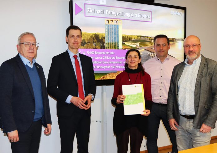 Fünf Personen stehen nebeneinander in einem Besprechungsraum. In der Mitte hält eine Frau einen Ordner mit Planunterlagen in den Händen. Hinter der Gruppe ist ein Bildschirm mit einer Visualisierung des künftigen Scheibe-Sees und dem Schriftzug Ziel nach Fertigstellung des Reviers zu sehen. Die Personen lächeln in die Kamera und posieren zur Übergabe eines Genehmigungsbescheids.