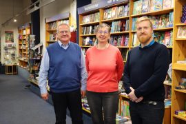 Drei Personen stehen in einer Buchhandlung vor gut gefüllten Bücherregalen. Links steht der Oberbürgermeister der Stadt Hoyerswerda, in der Mitte die Inhaberin des Buch und Musikhauses Sygusch, rechts der Wirtschaftsförderer der Stadt. Alle blicken in die Kamera.