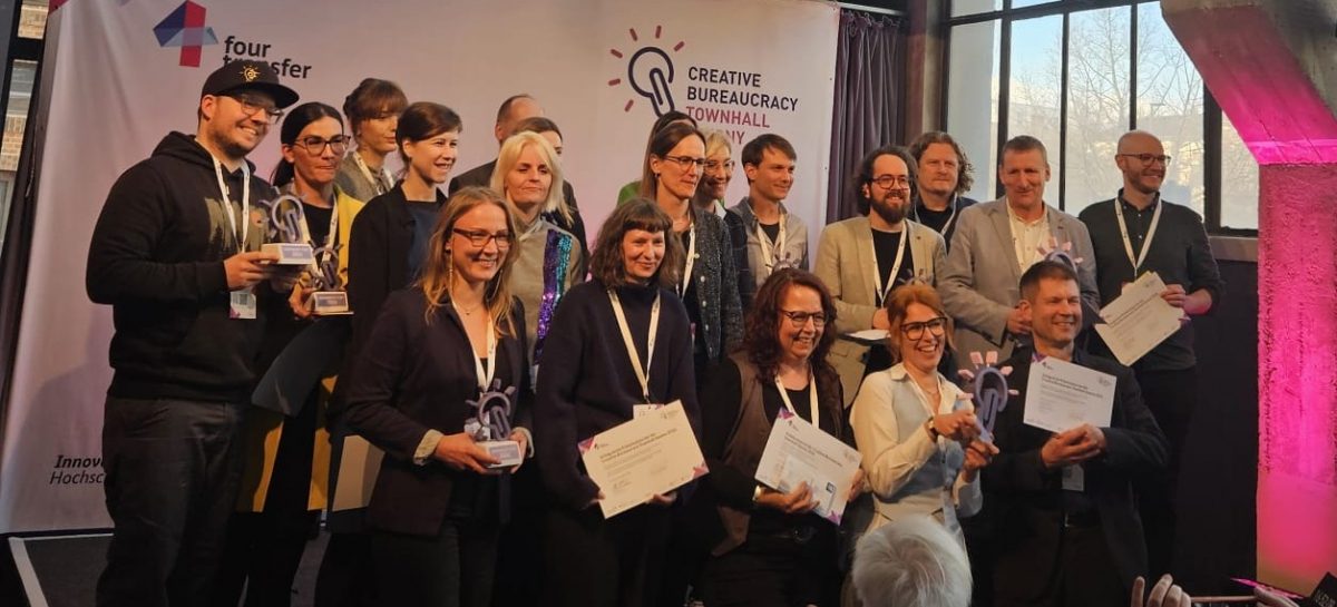 Eine Gruppe von etwa zwanzig Personen steht und kniet auf einer Bühne, einige halten Urkunden und gläserne Trophäen. Auf der Videowand darüber steht „Danke an alle Beteiligten!" Zuschauer im Vordergrund applaudieren und fotografieren.