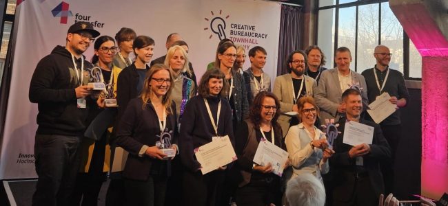 Eine Gruppe von etwa zwanzig Personen steht und kniet auf einer Bühne, einige halten Urkunden und gläserne Trophäen. Auf der Videowand darüber steht „Danke an alle Beteiligten!" Zuschauer im Vordergrund applaudieren und fotografieren.