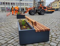 Eine moderne Pflanzbank mit Holzsitzfläche und bepflanztem Kasten steht auf einem gepflasterten Marktplatz. Im Hintergrund arbeiten Mitarbeitende in orangefarbener Arbeitskleidung mit einem Radlader an der Aufstellung weiterer Elemente.