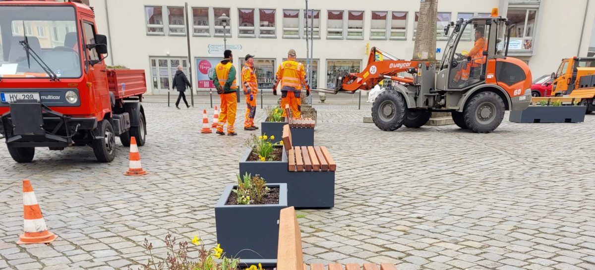 Mehrere moderne Pflanzbänke und Pflanzkübel stehen auf dem gepflasterten Marktplatz. Mitarbeitende des Regiebetriebs Baubetriebshof und Stadtgrün in orangefarbener Arbeitskleidung stellen die Elemente mit einem Radlader auf und bepflanzen sie.