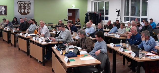 Mehrere Personen sitzen in einem Sitzungssaal an Tischen und nehmen an einer Stadtratssitzung teil. Vor ihnen liegen Unterlagen, Tablets und Mikrofone. Im Hintergrund sind weitere Teilnehmende sowie eine Kamera für den Livestream zu sehen. An der Wand hängt das Stadtwappen.