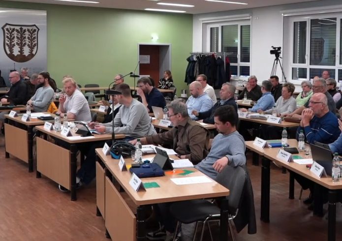 Mehrere Personen sitzen in einem Sitzungssaal an Tischen und nehmen an einer Stadtratssitzung teil. Vor ihnen liegen Unterlagen, Tablets und Mikrofone. Im Hintergrund sind weitere Teilnehmende sowie eine Kamera für den Livestream zu sehen. An der Wand hängt das Stadtwappen.