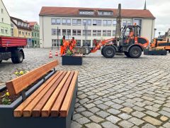 Mehrere moderne Pflanzbänke und Pflanzkübel stehen auf dem gepflasterten Marktplatz. Mitarbeitende des Regiebetriebs Baubetriebshof und Stadtgrün in orangefarbener Arbeitskleidung stellen die Elemente mit einem Radlader auf und bepflanzen sie.
