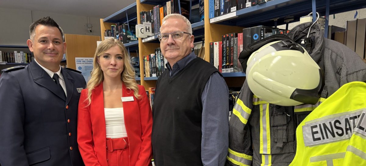 Drei Personen stehen in der Stadtbibliothek „Brigitte Reimann“ vor einem Bücherregal. Zu sehen sind Oberbürgermeister Torsten Ruban-Zeh, Benny Bastisch von der Feuerwehr Hoyerswerda sowie Sara Reimann. Rechts im Bild hängen Feuerwehrschutzkleidung und ein Feuerwehrhelm.