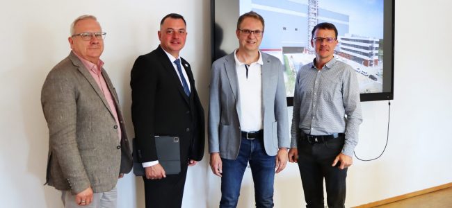 Vier Männer stehen nebeneinander in einem hellen Raum vor einem großen Bildschirm. Von links nach rechts: Torsten Ruban-Zeh (Oberbürgermeister Stadt Hoyerswerda), Udo Witschas (Landrat Landkreis Bautzen), Prof. Dr.-Ing. Günther Prokop (TU Dresden, Sprecher des Smart Mobility Lab) und Tobias Zschieschick (Koordinator Smart Mobility Lab). Auf dem Bildschirm ist eine Visualisierung eines modernen Gebäudekomplexes zu sehen.