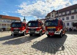 Drei neue rote Löschgruppenfahrzeuge vom Typ LF 10 stehen nebeneinander auf dem Marktplatz in Hoyerswerdas Altstadt. Im Hintergrund sind historische Gebäude und Kirchturmspitzen unter einem teilweise bewölkten Himmel zu sehen.