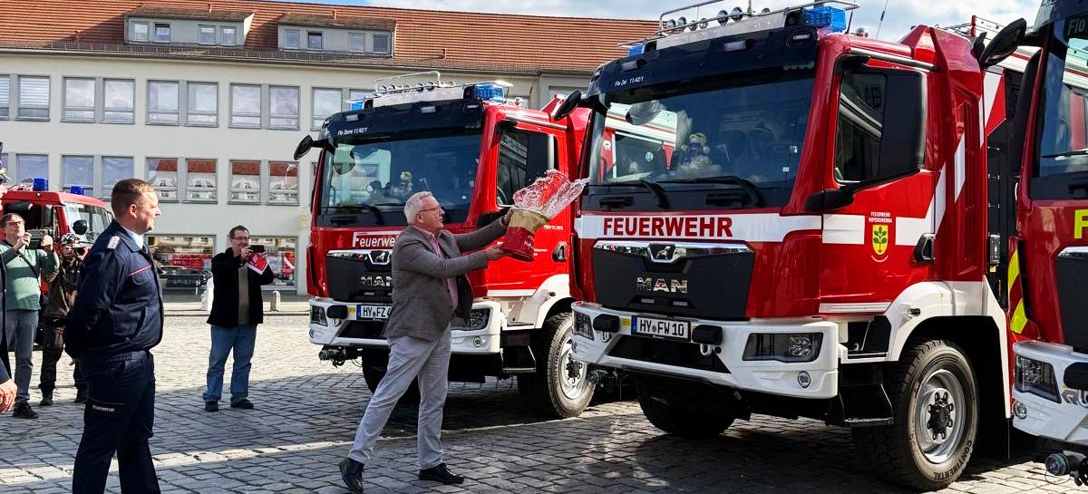Ein Mann tauft vor mehreren Feuerwehrfahrzeugen ein neues Löschgruppenfahrzeug mit einer Flasche. Daneben stehen Feuerwehrangehörige und Gäste der offiziellen Fahrzeugübergabe auf dem Marktplatz in Hoyerswerda.