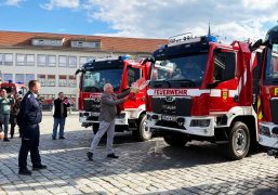 Ein Mann tauft vor mehreren Feuerwehrfahrzeugen ein neues Löschgruppenfahrzeug mit einer Flasche. Daneben stehen Feuerwehrangehörige und Gäste der offiziellen Fahrzeugübergabe auf dem Marktplatz in Hoyerswerda.