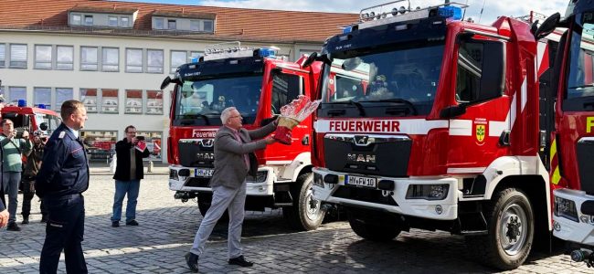 Ein Mann tauft vor mehreren Feuerwehrfahrzeugen ein neues Löschgruppenfahrzeug mit einer Flasche. Daneben stehen Feuerwehrangehörige und Gäste der offiziellen Fahrzeugübergabe auf dem Marktplatz in Hoyerswerda.