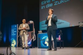 Auf der Bühne der Lausitzhalle Hoyerswerda stehen Oberbürgermeister Torsten Ruban-Zeh und Sachsens Kultusminister Conrad Clemens bei der Eröffnung der DLR-Raumfahrt-Show. Im Hintergrund sind Bühnenelemente und eine große Projektion mit dem Titel „Eine Zeitreise in die Zukunft“ zu sehen.