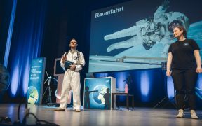 Zwei Mitarbeitende der DLR-Raumfahrt-Show stehen auf der Bühne der Lausitzhalle Hoyerswerda. Im Hintergrund ist auf einer großen Leinwand ein Astronaut im Weltraum mit der Überschrift „Raumfahrt“ zu sehen. Einer der Moderatoren trägt einen Raumanzug.