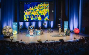 Auf einer großen Leinwand in der Lausitzhalle Hoyerswerda ist während der DLR-Raumfahrt-Show eine Wärmebildaufnahme des Publikums zu sehen. Die Bühne ist mit verschiedenen Experimentierstationen und Technikmodellen aufgebaut.