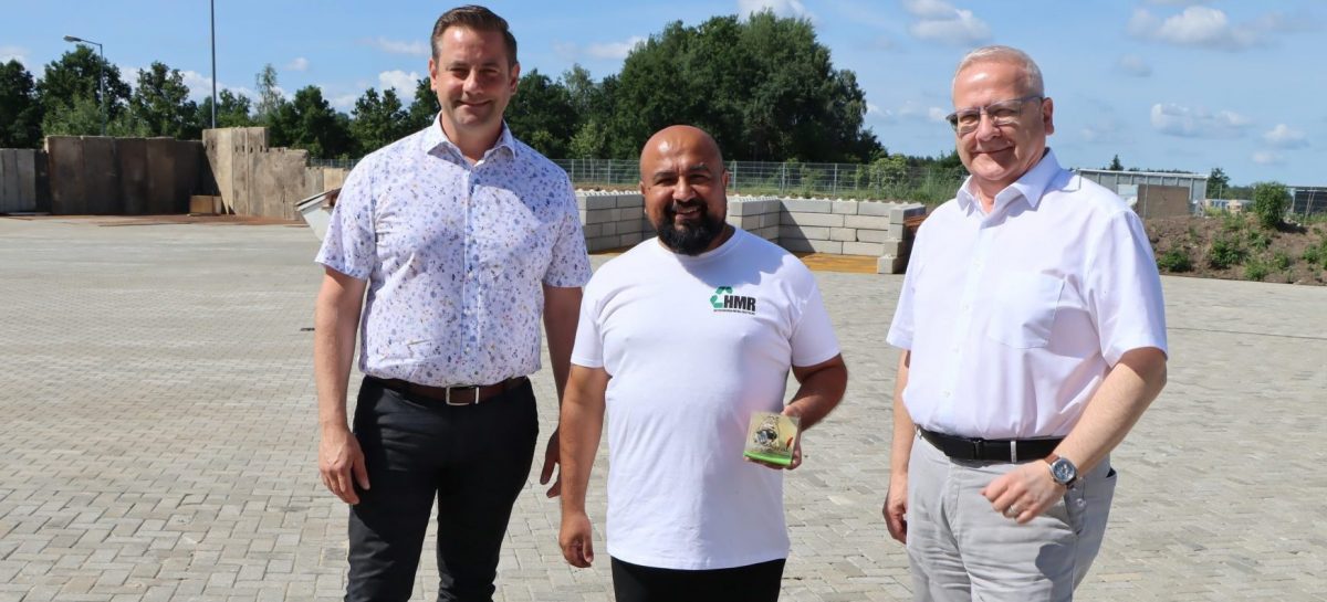 Drei Männer stehen auf dem Betriebsgelände der Hoyerswerda Metall Recycling GmbH im Gewerbegebiet Nardt. Von links nach rechts: Christian Hoffmann, Geschäftsführer Hajrudin Hidanovic und Oberbürgermeister Torsten Ruban-Zeh. Hajrudin Hidanovic hält ein kleines Präsent in den Händen. Im Hintergrund sind die befestigten Betriebsflächen des neu eröffneten Recyclingunternehmens zu sehen.
