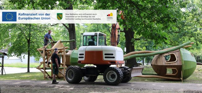Mitarbeiter montieren mit Unterstützung eines Baggers ein großes Holzspielgerät auf dem Gelände des neuen Spielplatzes im Altstadtpark.