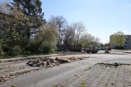 Blick auf das ehemalige Busbahnhofareal im Wohnkomplex VI vor Beginn der Bauarbeiten. Im Vordergrund befinden sich befestigte Flächen und Rückbauarbeiten, im Hintergrund ist die erhaltene DDR-Formsteinwand zwischen Bäumen und Sträuchern zu erkennen.