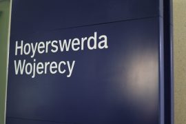 Blaues Bahnhofsschild mit der zweisprachigen Beschriftung „Hoyerswerda/Wojerecy“ in deutscher und sorbischer Sprache im Bahnhofsgebäude von Hoyerswerda.