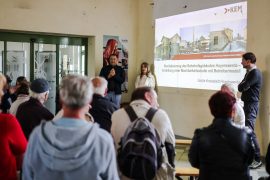 Teilnehmende eines Bürgerworkshops sitzen in der Bahnhofshalle Hoyerswerda und verfolgen eine Präsentation zur Machbarkeitsstudie für das Bahnhofsgebäude. Vor einer projizierten Präsentationsfolie stehen Vertreterinnen und Vertreter der Stadt sowie der KEM.