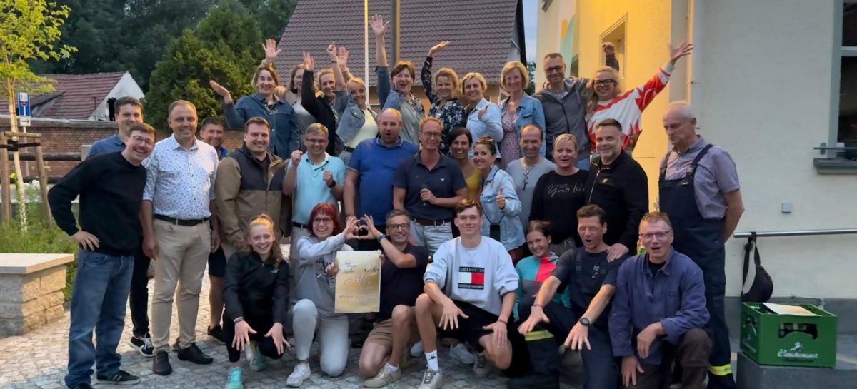Gruppenfoto mit Bürgermeister Mirko Pink und rund 30 Engagierten aus Dörgenhausen. Die Personen stehen und knien vor einem Gebäude, viele lächeln, winken oder zeigen Jubelgesten. In der Mitte wird ein Schild zur Auszeichnung beim Bundeswettbewerb „Unser Dorf hat Zukunft“ gehalten.