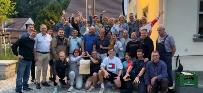 Gruppenfoto mit Bürgermeister Mirko Pink und rund 30 Engagierten aus Dörgenhausen. Die Personen stehen und knien vor einem Gebäude, viele lächeln, winken oder zeigen Jubelgesten. In der Mitte wird ein Schild zur Auszeichnung beim Bundeswettbewerb „Unser Dorf hat Zukunft“ gehalten.