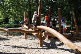 Mehrere Kinder klettern auf einer großen Holz-Kletteranlage des neuen Spielplatzes „Baumschätze“ im Altstadtpark unter schattigen Bäumen.